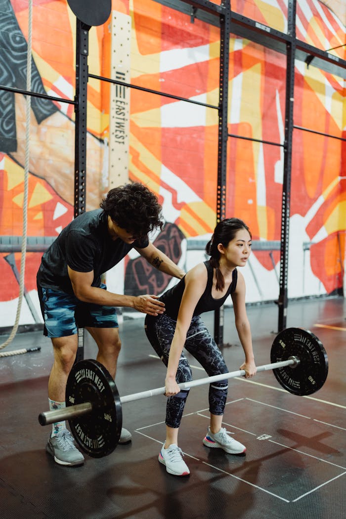 why-choose-us A young woman lifting a barbell in a gym with a trainers guidance. Perfect for fitness and training themes.