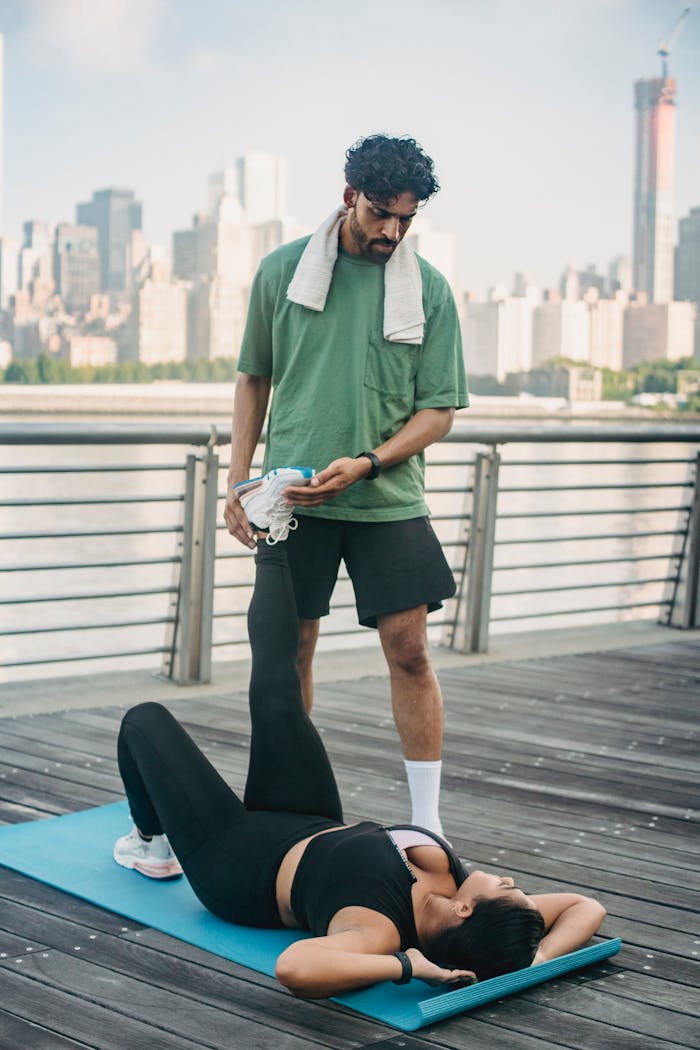 Services-03 A man helps a woman stretch on a yoga mat by the waterfront with city skyline backdrop.